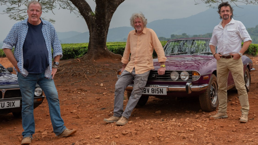 Jeremy Clarkson, James May and Richard Hammond filming the final The Grand Tour series on Amazon. They are stood together by cars on a dusty/clay surface outdoors with a large tree behind them. They're all wearing causal clothing.