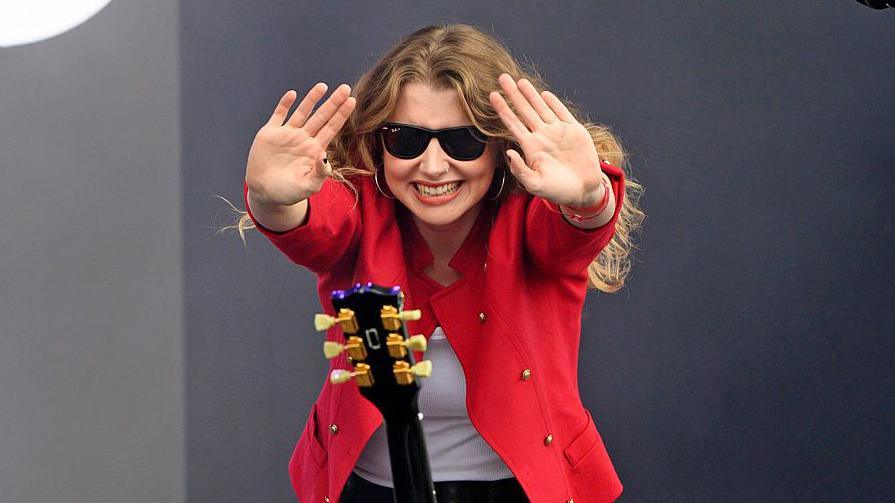 Ellur - Ella McNamara - waving at the crowd wearing sunglasses, with a guitar in front of her. She is wearing a red jacket and a white T-shirt. 
