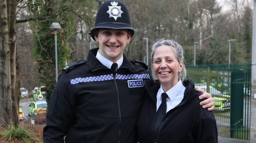 Charlie Bone with his mother Victoria at a police building. He is wearing a police helmet and jacket with a police badge, she is wearing a half-zipped black jacket, white shirt and black tie. 