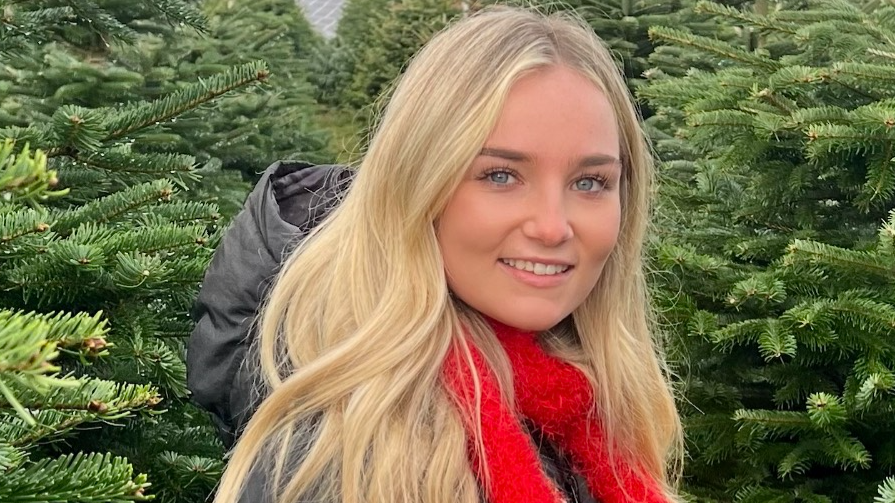 Emily is standing in a grove of Christmas trees. she is wearing a grey parka and has a red scarf tied at her neck. She has long wavy blonde hair, blue eyes and is wearing light makeup. 