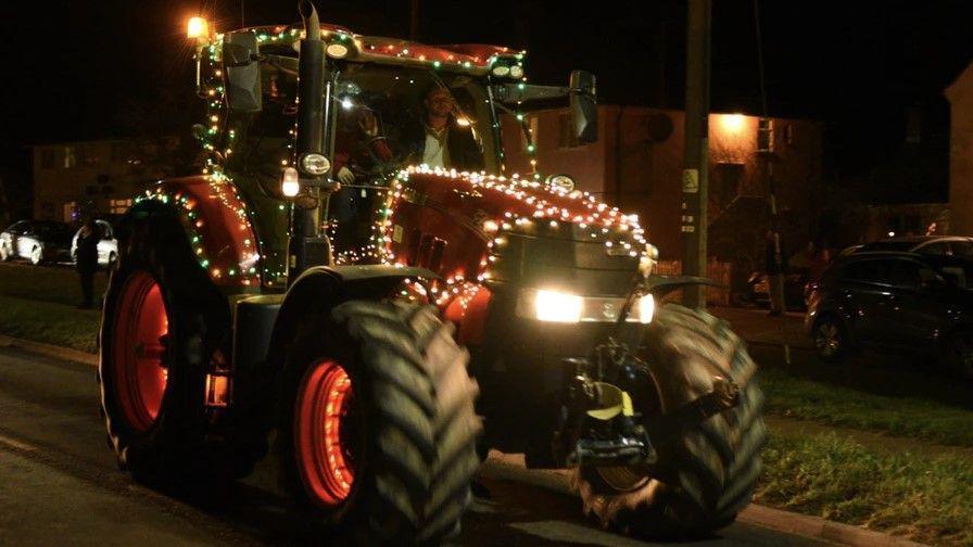 A red tractor decorated with white lights is travelling through a darkly lit village street  