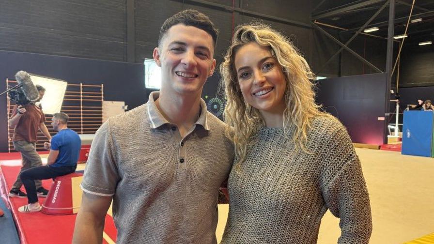 A man wearing a grey polo shirt stood in a gym next to a blonde woman with blonde hair. They are both smiling at the camera 