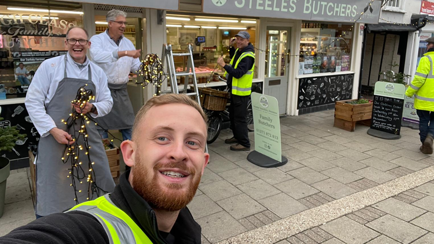 Jack Coleman, who has short fair hair and a beard, smiles in this selfie with butchers and workmen putting up lights on the front of a butcher's shop.