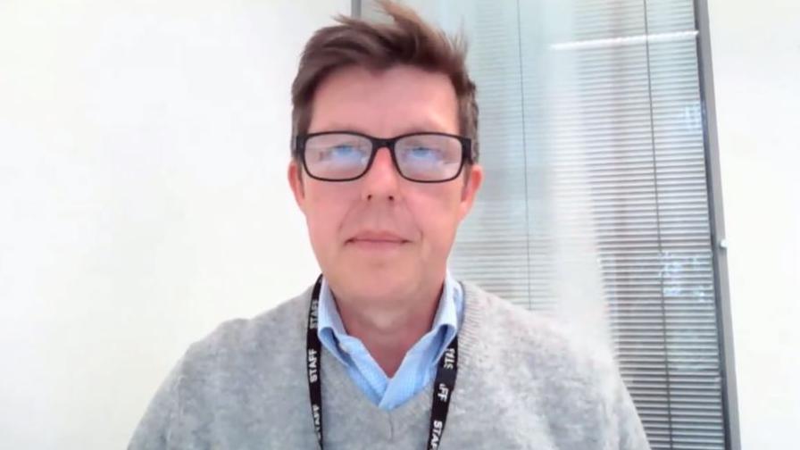 Rory Sanderson has light brown hair and wears black rimmed glasses. He has a staff lanyard around his neck and is wearing a grey pullover and a blue shirt, which is open at the neck.