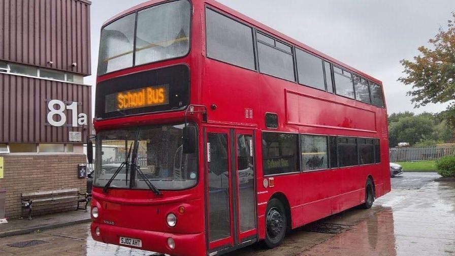 A red double-decker bus stands in school grounds. A sign at the front reads "School Bus". It has been raining and there are puddles on the ground.