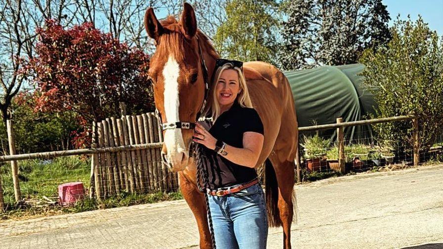 A blonde white woman with black sunglasses over her head wearing a black shirt and blue jeans is standing next to and holding a brown horse with a white patch on its nose.