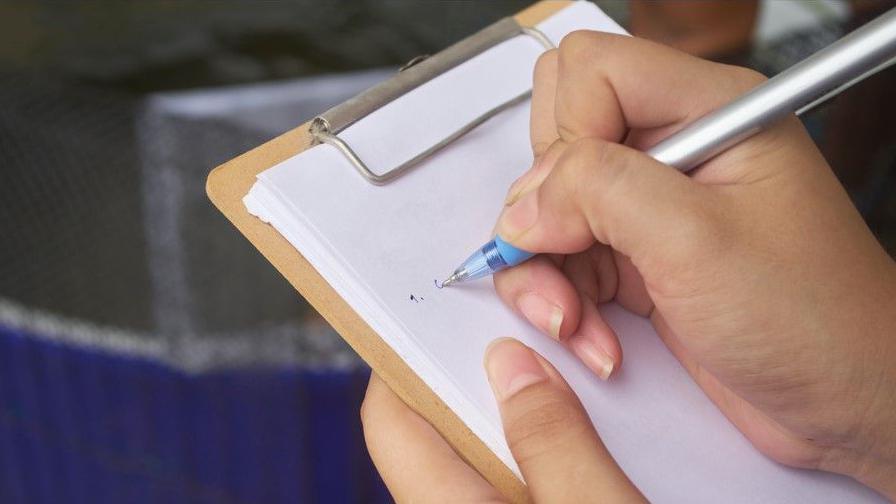 An image of a woman with a pen in her right hand, writing on a clipboard.
