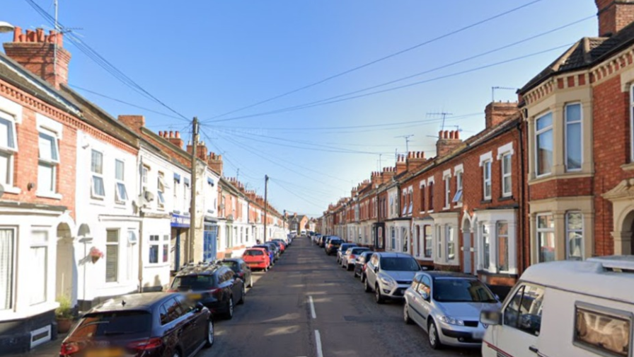 A narrow street full of Victorian terraced houses with many cars parallel parked. 