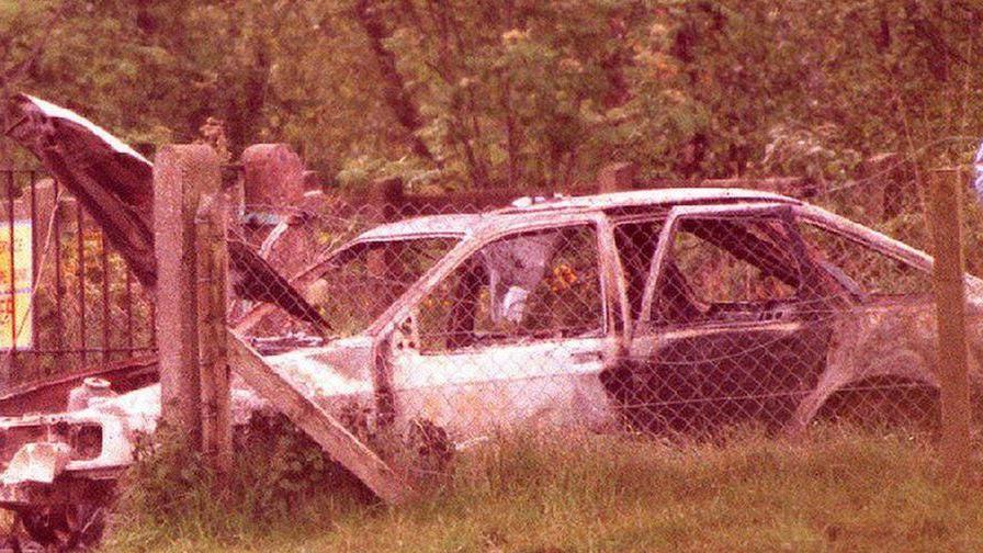 Old image of a burnt out white car behind a wired fence