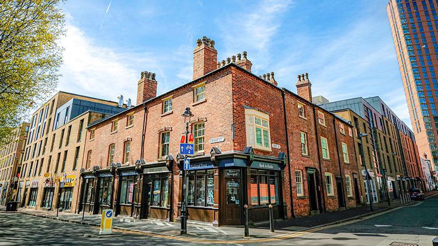 The corner of Hurst Street in Birmingham, featuring rows of shops. The buildings include dozens of windows and shop fronts, with street signs and a car seen in the background.