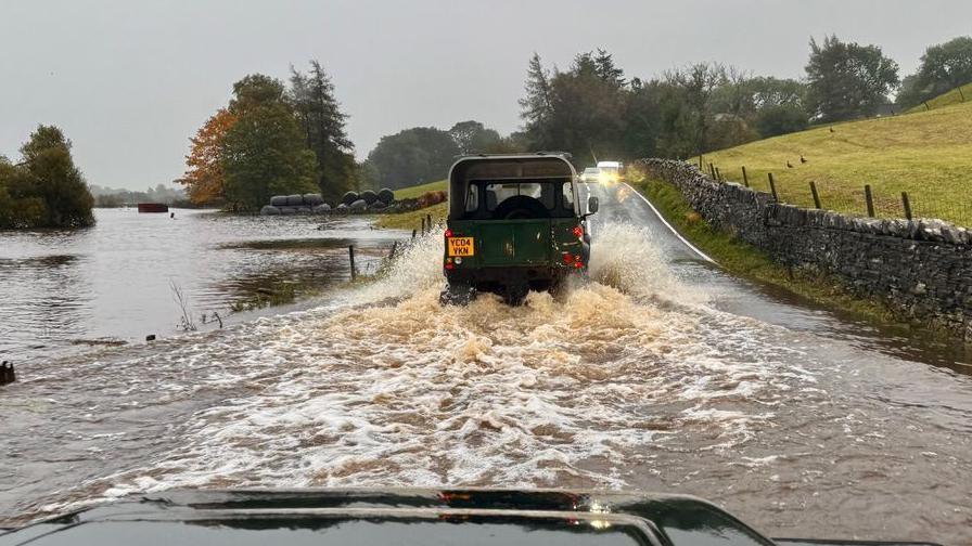 A flooded rural road viewed from inside a vehicle, with another off-road vehicle ahead driving through deep water. The road is partially submerged due to overflow from a nearby river. On the right, a stone wall borders a grassy hillside; on the left, trees and floodwater stretch into the distance. The sky is overcast.