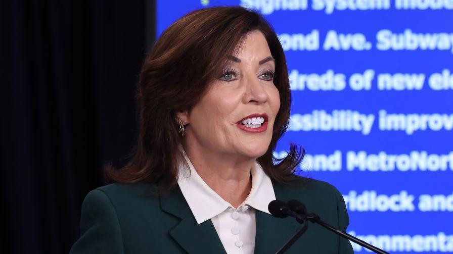 New York Governor Kathy Hochul wears a white shirt and dark jacket. She speaks into a microphone while standing in front of a blue and white wall