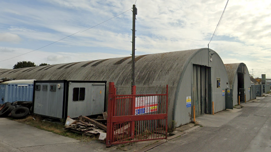 Two buildings on an industral estate. The buildings are tube shaped and grey. There is a red fence in the foreground and an electricity pylon.