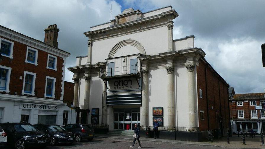 The Orion Cinema in Dereham. It has a white facade with columns at the front. There is a sign that says "Orion Cinema" above the entrance doors. The side of the building is made of red brick. The cinema is based in a town square.