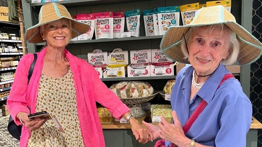 Susan (left) in a pink jacket and floral dress with a straw hat on. She looks like she may be in her 60s
Her mum, who is noticeably owner, is wearing a blue/purple blouse and also a straw hat. The pair are holding hands and posing for a photo from the inside of a store.