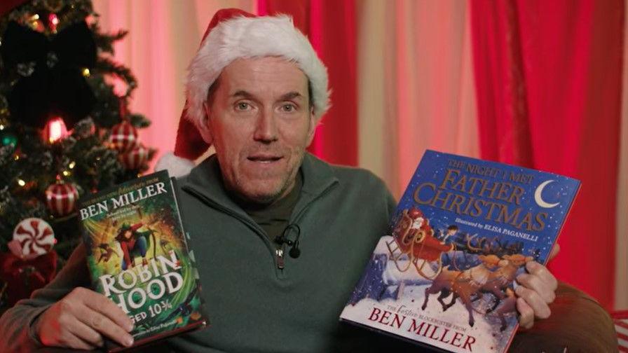 Ben Miller, wearing a Santa hat, is holding two of his books whilst sat next to a Christmas tree. One book is entitled Robin Hood aged 10 3/4 and the other book is called The Night I Met Father Christmas.