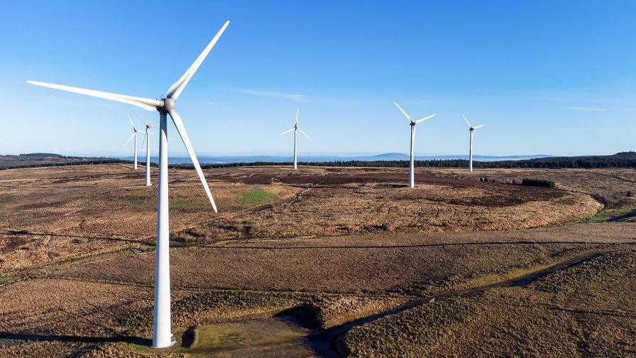 A wind farm in Northern Ireland, on farm land in front of a blue sky. There are multiple wind turbines in motion. 