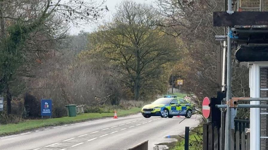 A road surrounded by trees with a police car parked on the road cordoning it off.