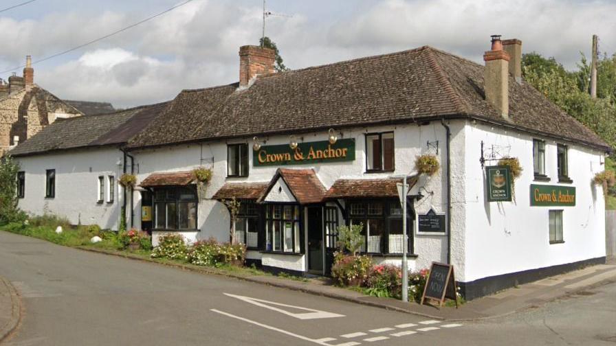 A picture of the Crown & Anchor pub. It is a white building on a corner with plants in front of it. The windows have black borders . The name of the pub is written in yellow with a green background.