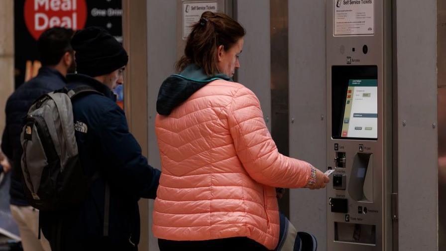 Woman dressed in a pink puffer jacket purchasing a train ticket from a self-service machine