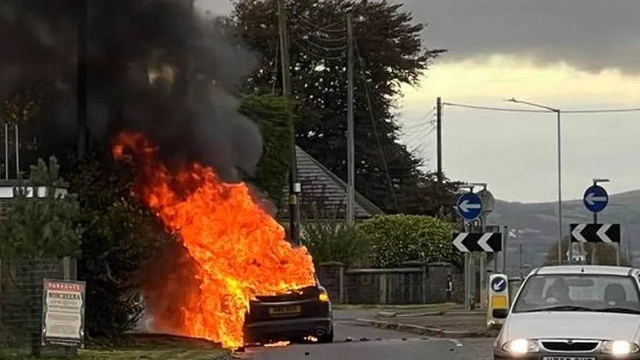 Huge amounts of fire and smoke stream from a burning car on the side of a road in the evening. Another car with headlights on drives past. The car is pulled over on a grass verge near a roundabout.