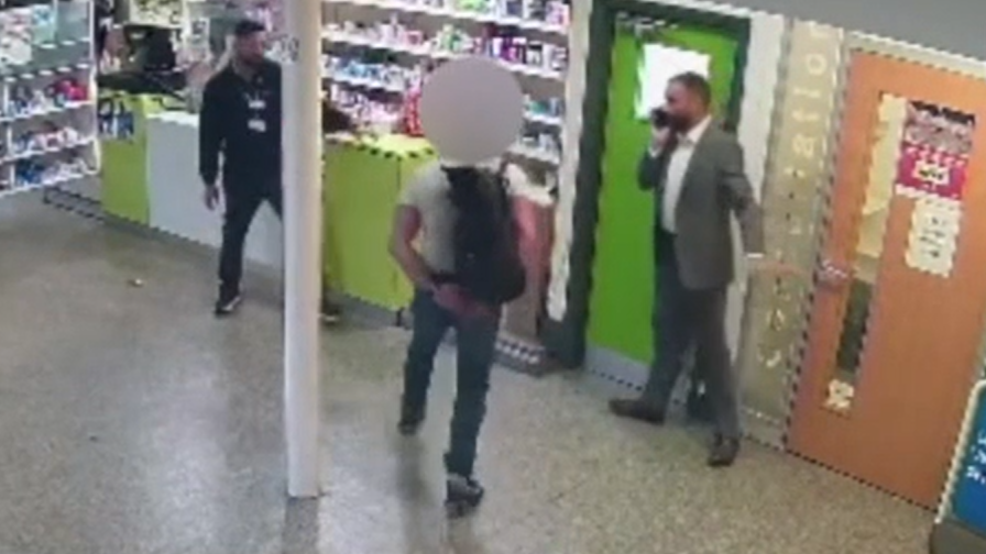 CCTV still of a man with a blurred-out face confronting two other men on the shop floor of a pharmacy. The main subject is in the middle in a white T-shirt and wearing a black backpack. The staff member on the left is in a black top with a lanyard round his neck, while the one on the right is in a grey suit with a phone to his ear.