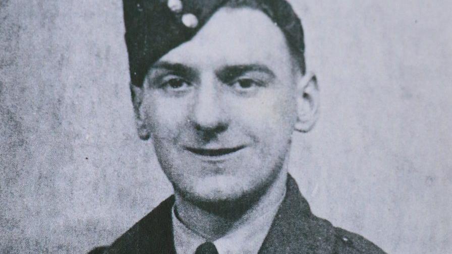 A black and white photo of a man in military uniform. He has a soft cap perched slightly on the right side of his head. He is clean shaven with dark hair and the top of a dark jacket can be seen over the white collar on a shirt.