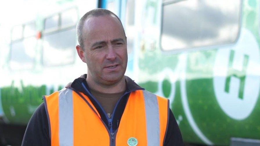 Wearing an orange hi-vis vest Stuart Hillmansen stands in front of a railway carriage.