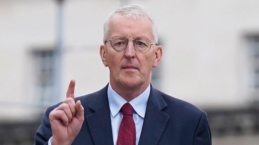 A man wearing a navy blue suit, white shirt and red tie. He is wearing spectacles and has his index finger raised.