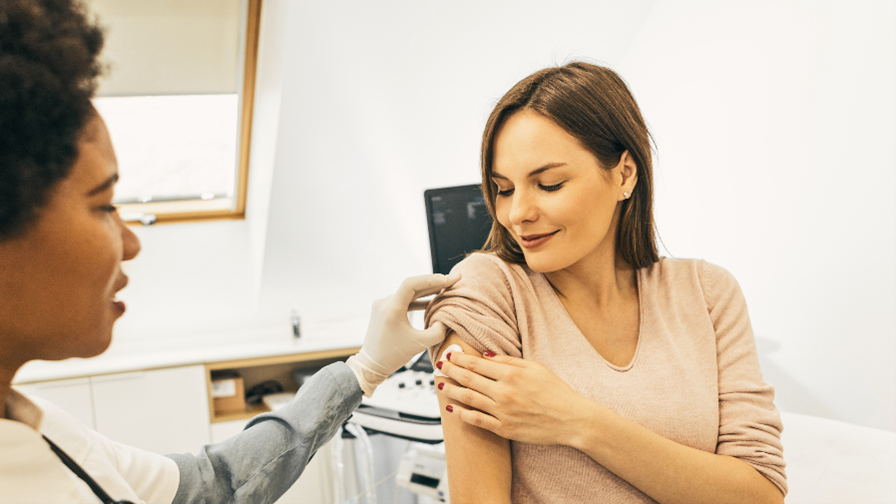 A doctor and a patient after a vaccine. The female patient is holding a cotton ball on her upper arm and looking down at it. The doctor is wearing latex gloves and holding the patient's sleeve up.