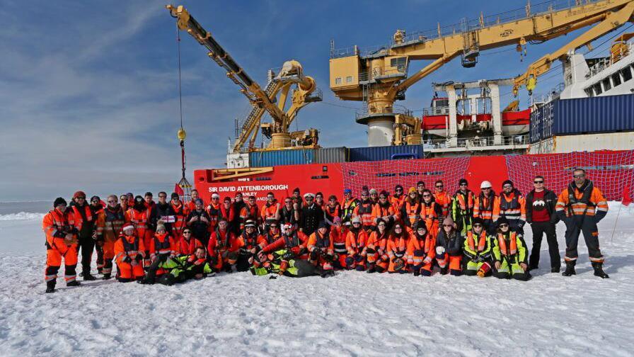 The crew of a ship are gathered on snow posing for the camera in front of the RRS Sir David Attenborough, which can be seen behind them