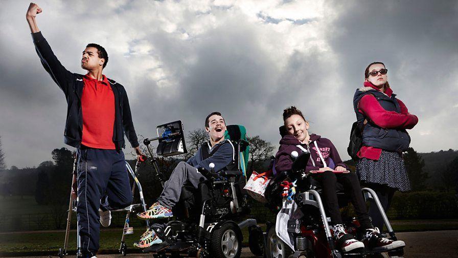 Four people outside, with one person on the left standing using sticks and raising a fist. Two others are seated in wheelchairs, one of which has a communication device attached, and another is stood with their arms crossed and wearing sunglasses. A cloudy sky and trees are visible in the background.