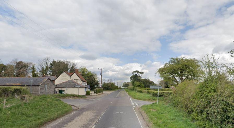 Road junction looking with a small lane running left to right there are road signs on the left side. To the left is a barn and other buildings.