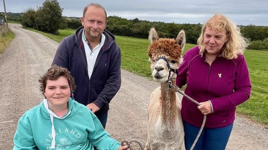 A smiling Emily is in a wheelchair and has mid-length hair with a braid. She is beside her father Andrew, who has short fair hair. Emily's mum is also stood next to them, with shoulder-length blonde hair. Both Emily and Caroline are holding a lead and are helping to guide an alpaca.