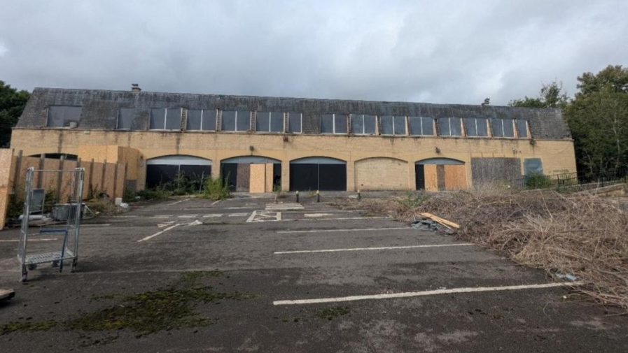An exterior view of the outside of the old co-op building. It is a wide one storey building with archways where windows used to be. Some of them are boarded up and there is a pile of old branches in the car park at the front of the building.