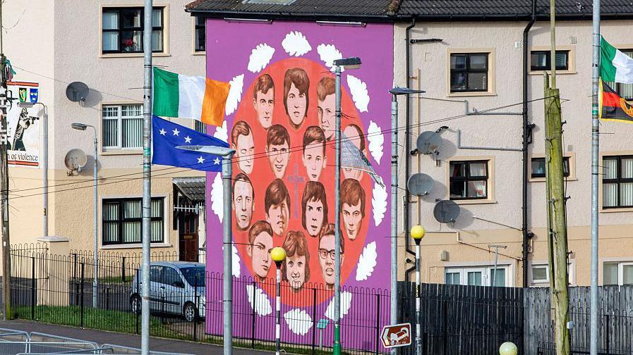 A purple mural at the end of a row of cream terrace houses which has the faces of the victims of Bloody Sunday. Their faces are in an orange circle on the mural. There is various flags hanging from the telegraph pole, included an Irish flag.
