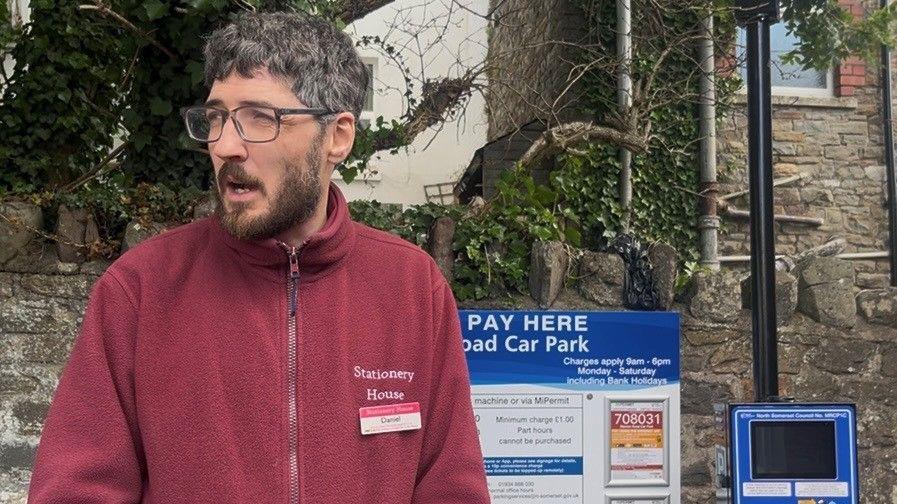 Daniel Adams at Marson Road Car Park, wearing a burgundy zip up fleece with Stationery House written on it. He is standing beside a parking meter and pay here sign.