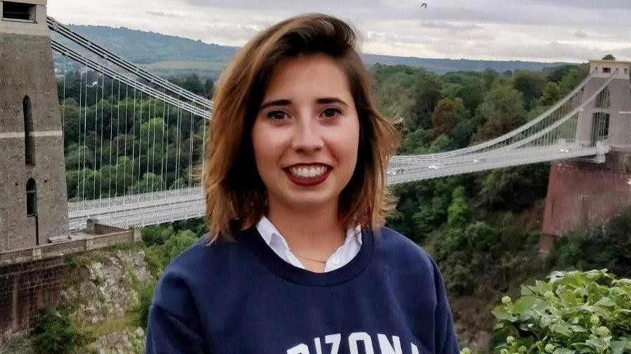 Marta Elena Vento stands with Clifton Suspension Bridge in the background. She has shoulder-length, brown hair and wears a navy blue sweatshirt, a white shirt and make-up.