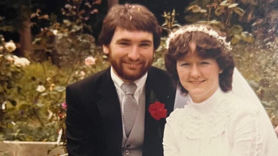 Afryl and Aled on their wedding day. Aled is on the left, smiling, and wearing a black suit with a silver waistcoat and tie. He has dark brown hair and a beard. Afryl is wearing a white wedding dress and veil. 