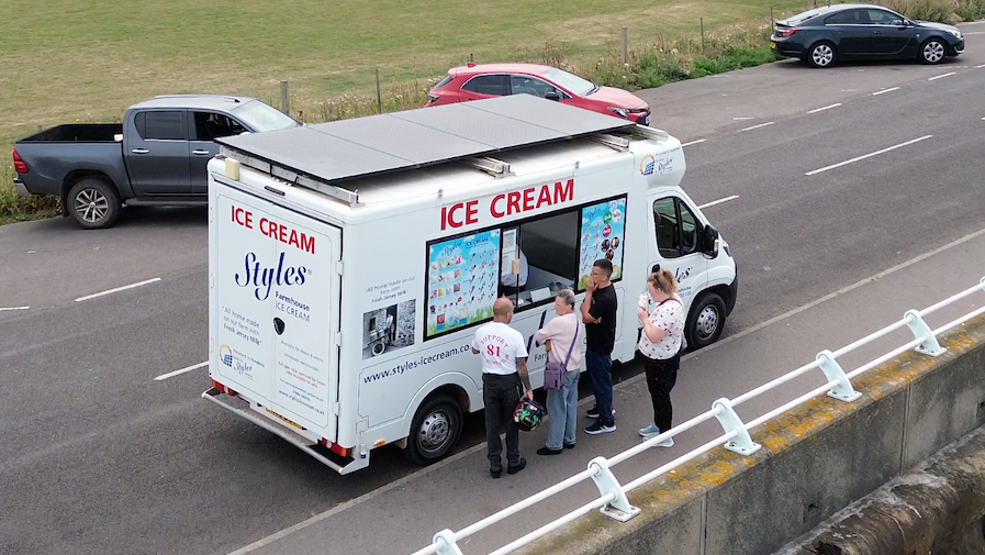 An ice cream van parked on a roadside, with four adults queueing outside. There are four solar panels attached to the van roof, covering it completely. The van has a picture menu next to a window hatch showing the ice creams it sells.