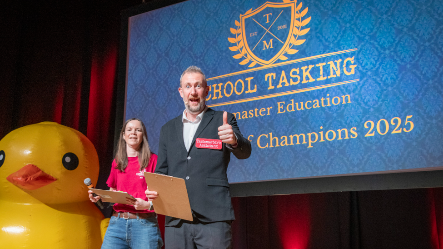 Comedian Alex Horne stands on a stage and looks at the camera doing a thumbs up. Next to him is a woman in a red shirt and blue jeans. On a screen behind them it says School Tasking and Champions 2025, although more text is obscured by Alex Horne. A very large yellow rubber duck is to the left of the picture.