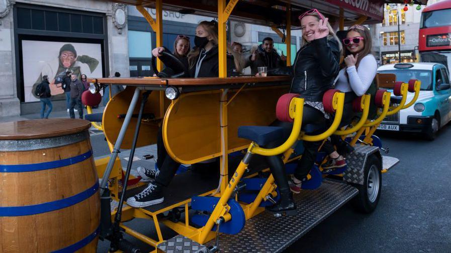 A party bike on Oxford Street with a group of women pedalling and smiling around a central bar table, surrounded pedestrians and road traffic.