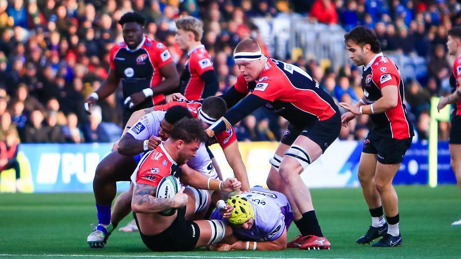 Action from Worcester Warriors v Cornish Pirates as a Pirates player is tackled to the ground still holding the ball and a Pirates lock prepares to jackle the second worcester defender.