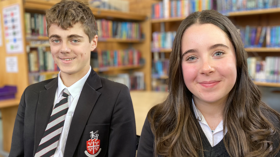 George and Emmy are sitting together. They're wearing the school uniform which is a black blazer and black jumper.  They are sitting in a library and there are shelves with books in the background