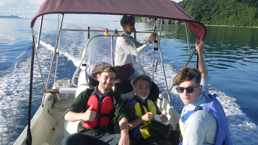 Four men are on an open boat with a small canopy. One is standing and driving the boat, the other three are seated. A young man with sunglasses is in the foreground, an elderly Japanese man with a white beard is in the centre with a man in his 20s sitting next to him. They are all wearing different colour life jackets