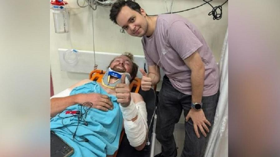 Harry Satloka pictured in the hospital bed with bandages, next to an unknown man doing a thumbs up 