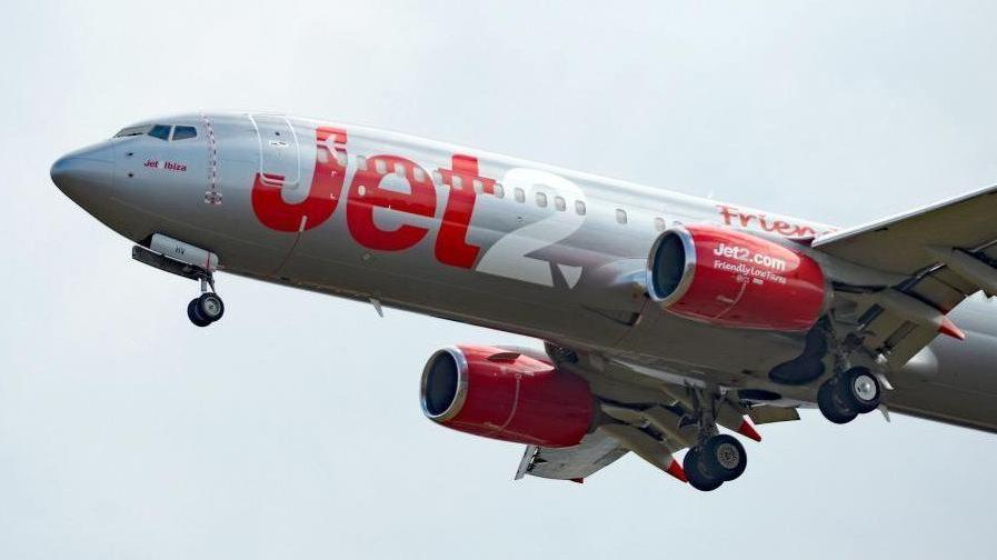 A Jet2 aeroplane flying against a pale grey sky. It is painted white and has Jet written in red and 2 written in white in large letters on its body.