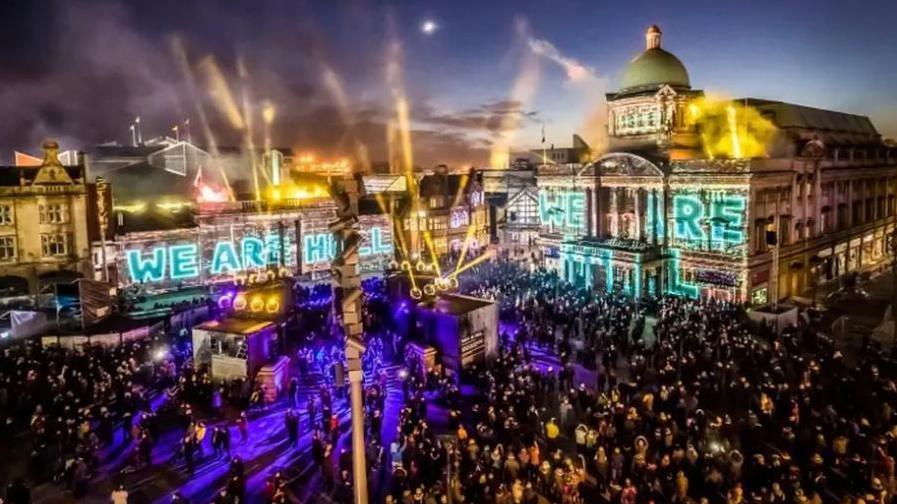 A city square is packed with people watching a spectacular light show at night in front of grand Edwardian buildings. The words 'WE ARE HULL' are projected on to the buildings, including one with a large dome.