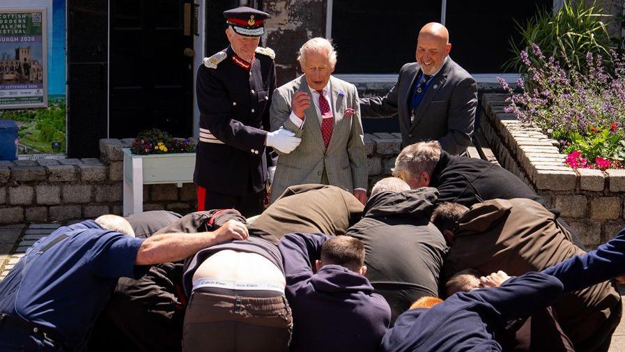 King Charles III is held back by a man in uniform and a man with a bald head as a group of men grapple for a ball in front of him