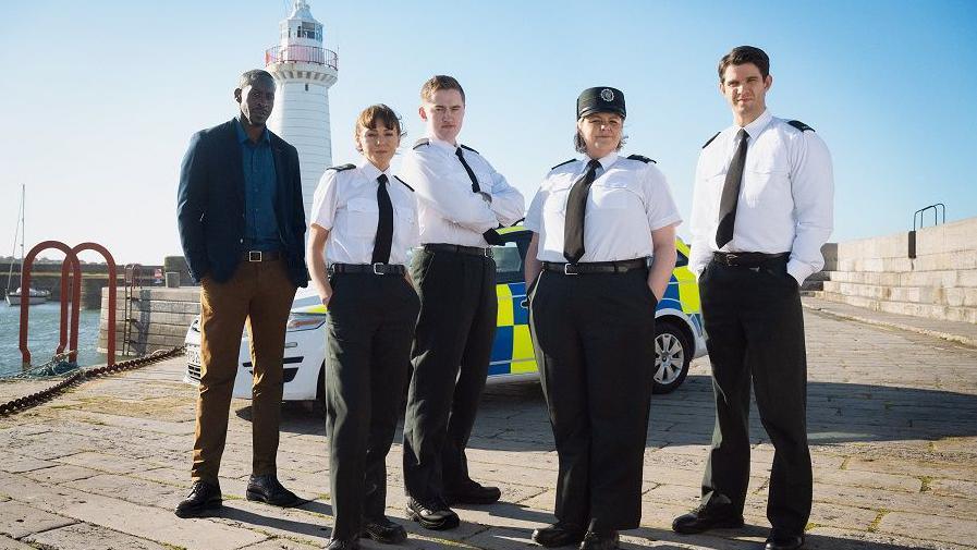 Five cast members of Hope Street, four of them in PSNI uniform stand in front of a police car on a pier with a lighthouse in the background